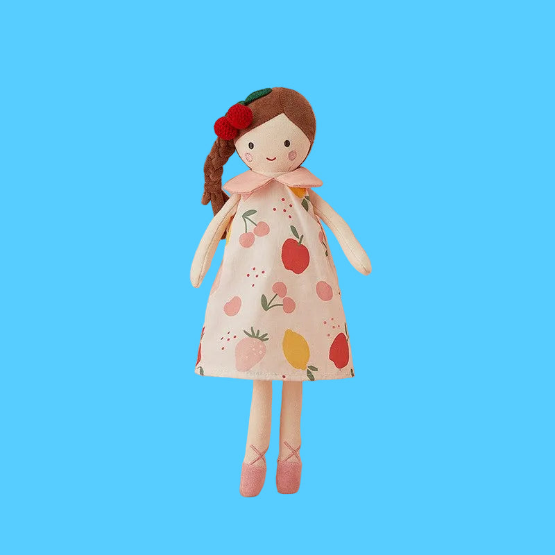 Boneca de pano artesanal com cabelo castanho em trança lateral, usando um prendedor de cereja na cabeça e um vestido estampado com frutas coloridas como morangos e maçãs, sobre fundo azul claro.
