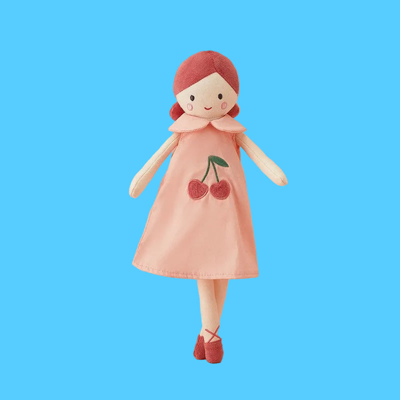 Boneca de pano artesanal com cabelo curto vermelho, usando um vestido simples na cor rosa salmão com estampa central de duas cerejas, sobre fundo azul claro.
